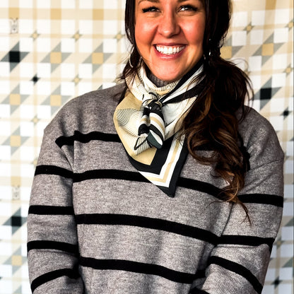 Woman wearing a gray striped sweater with black stripes, beige pants, and a patterned scarf against a geometric background.